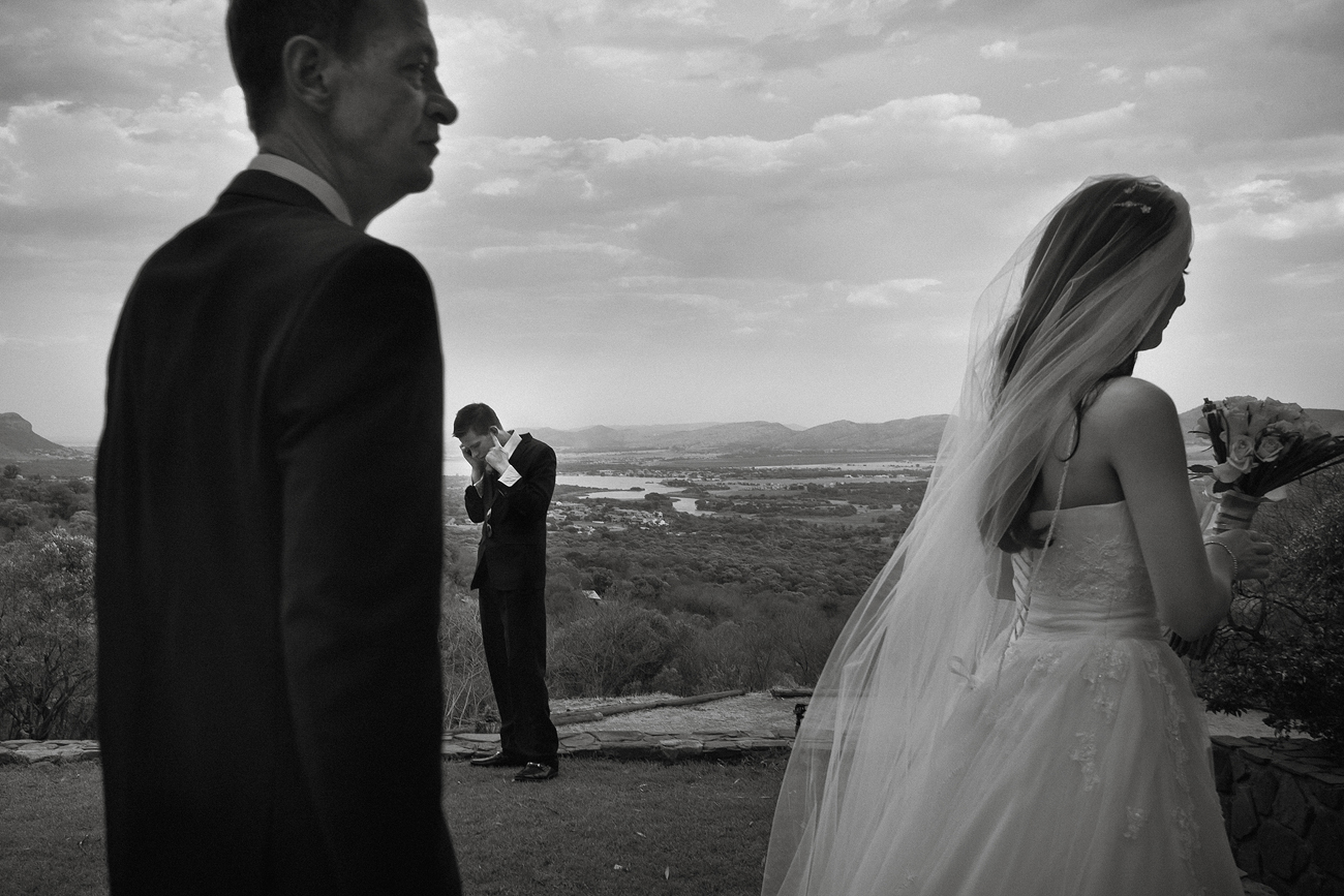 Documentary wedding photographer capturing a candid moment: groom on phone while bride and father walk in the foreground.