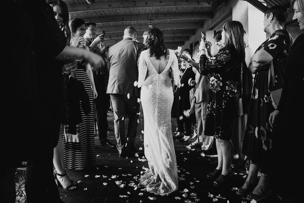 Bride, groom and confetti with guests framing and photographing them at Cradle Valley Guest Farm, taken in black and white and from behind.