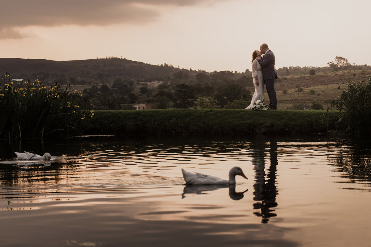 Bridal couple standing near the dam at Cradle Valley Guest House in Muldersdrift, with ducks gliding across the water in the foreground.